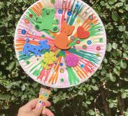 A colorful fan made out of a paper plate and decorated with stickers in front of foliage