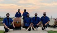 Members of Makoto Taiko pictured in uniform next to drums, against a sunset sky.