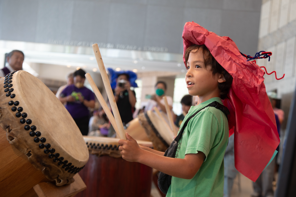 visitor wearing paper hat and p[laying taiko drum at family festival